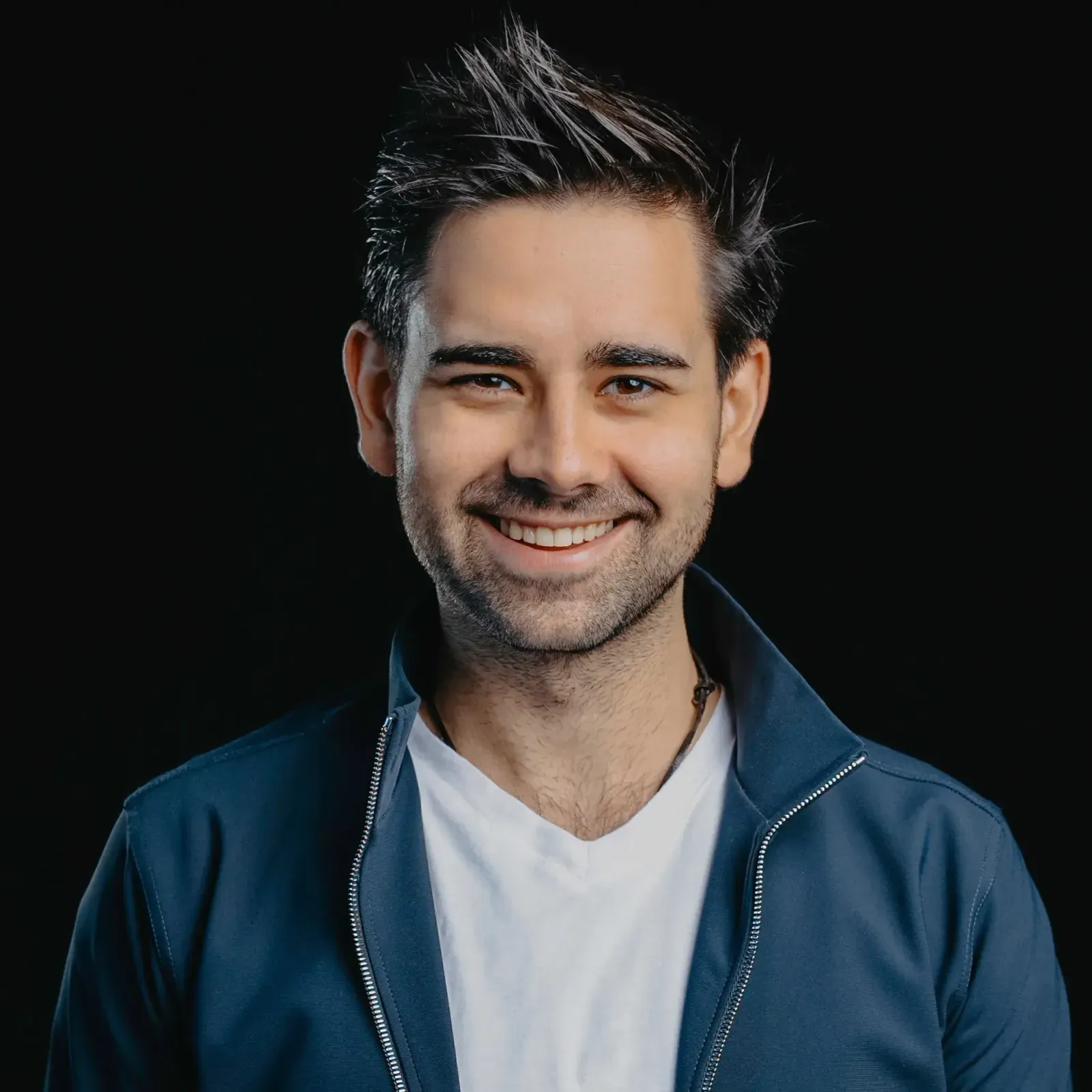 Smiling man with dark, upswept hair, white T-shirt and dark blue jacket in front of a black background.