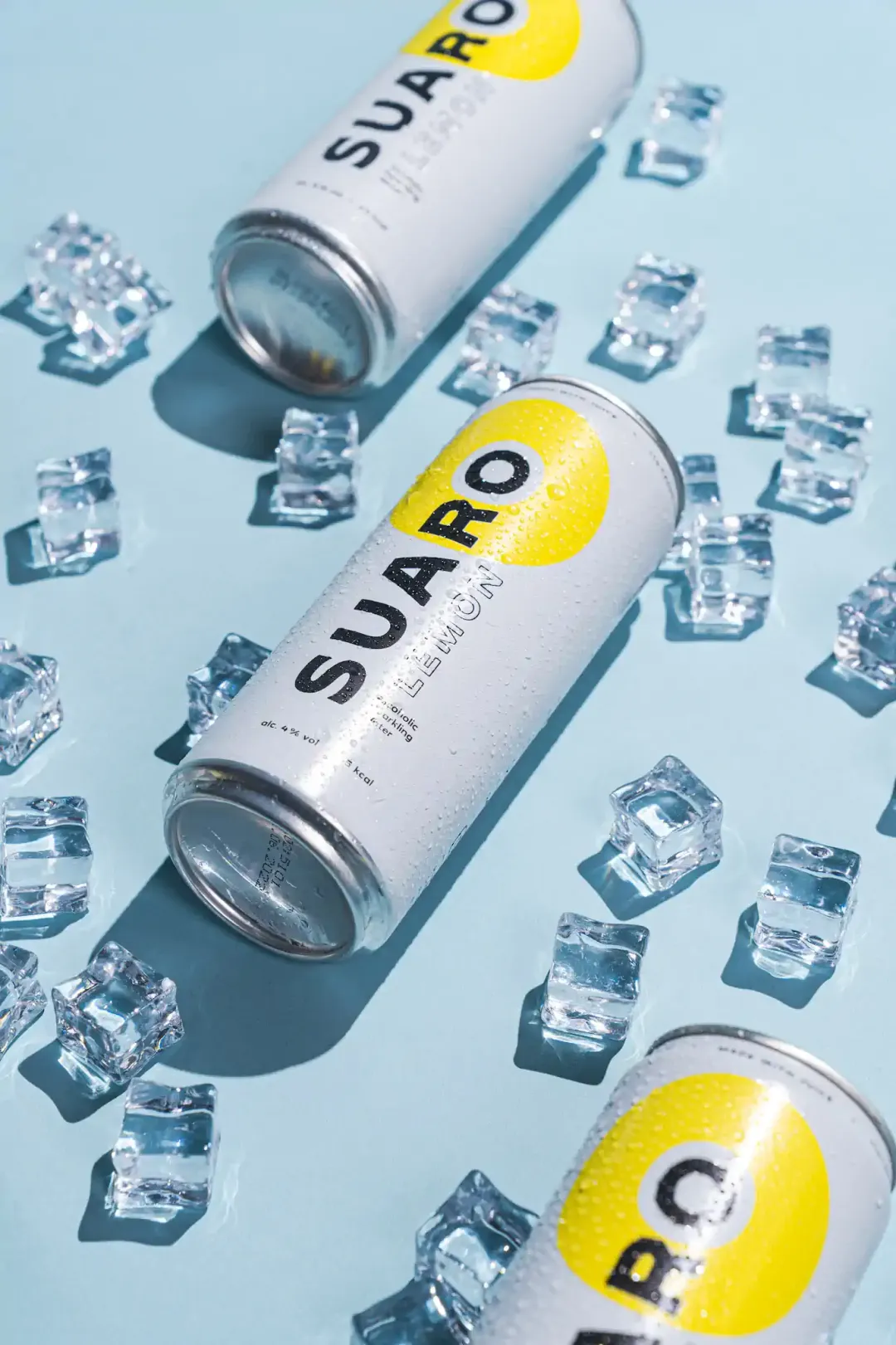 Zwei SUARO Lemon Dosen auf hellblauem Untergrund mit gestreuten Eiswürfeln – SUARO Getränkeshooting